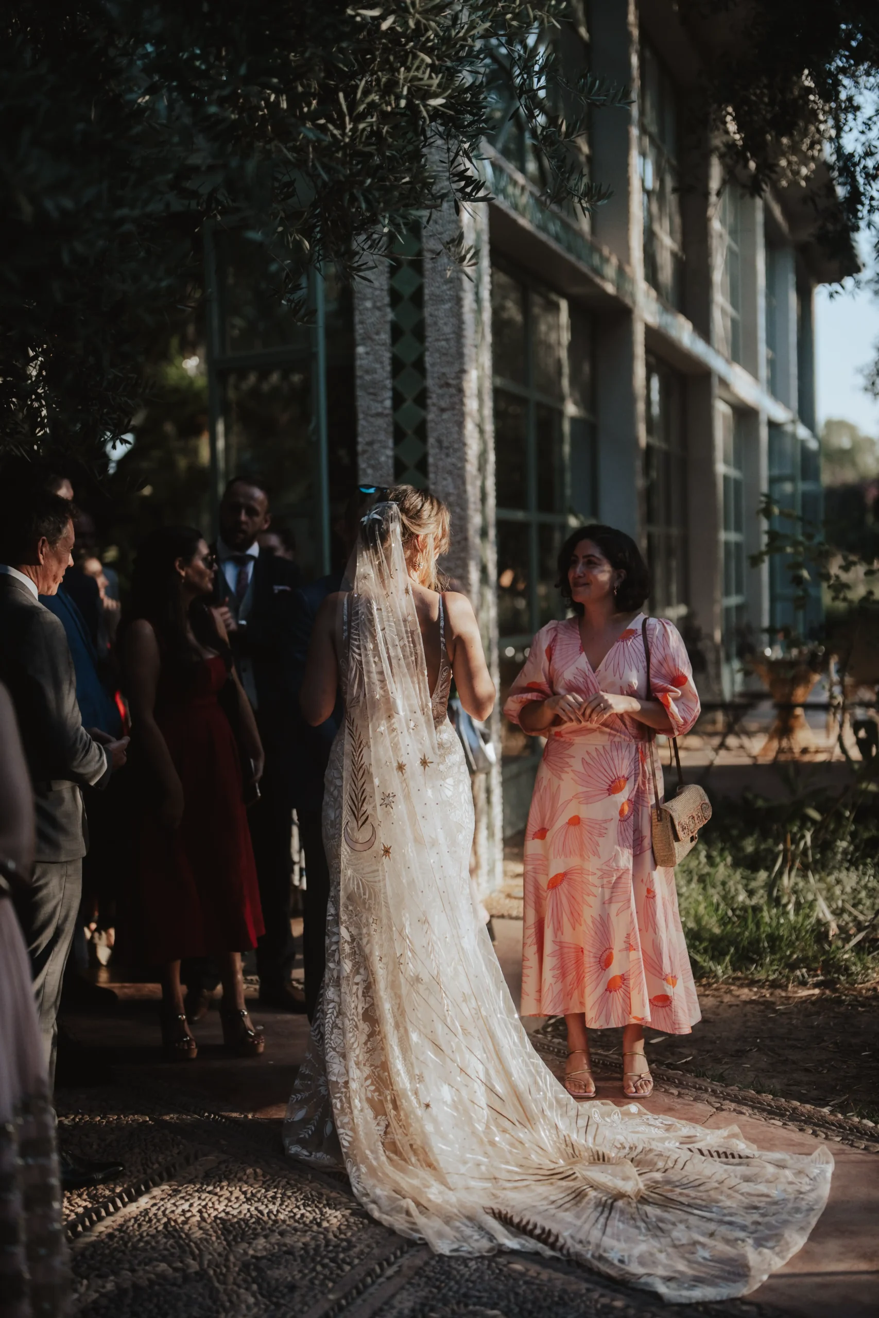 A bride in a dress with a long embroidered train talks to a woman in a pink floral dress at an outdoor wedding. Guests and a modern glass building are in the background.