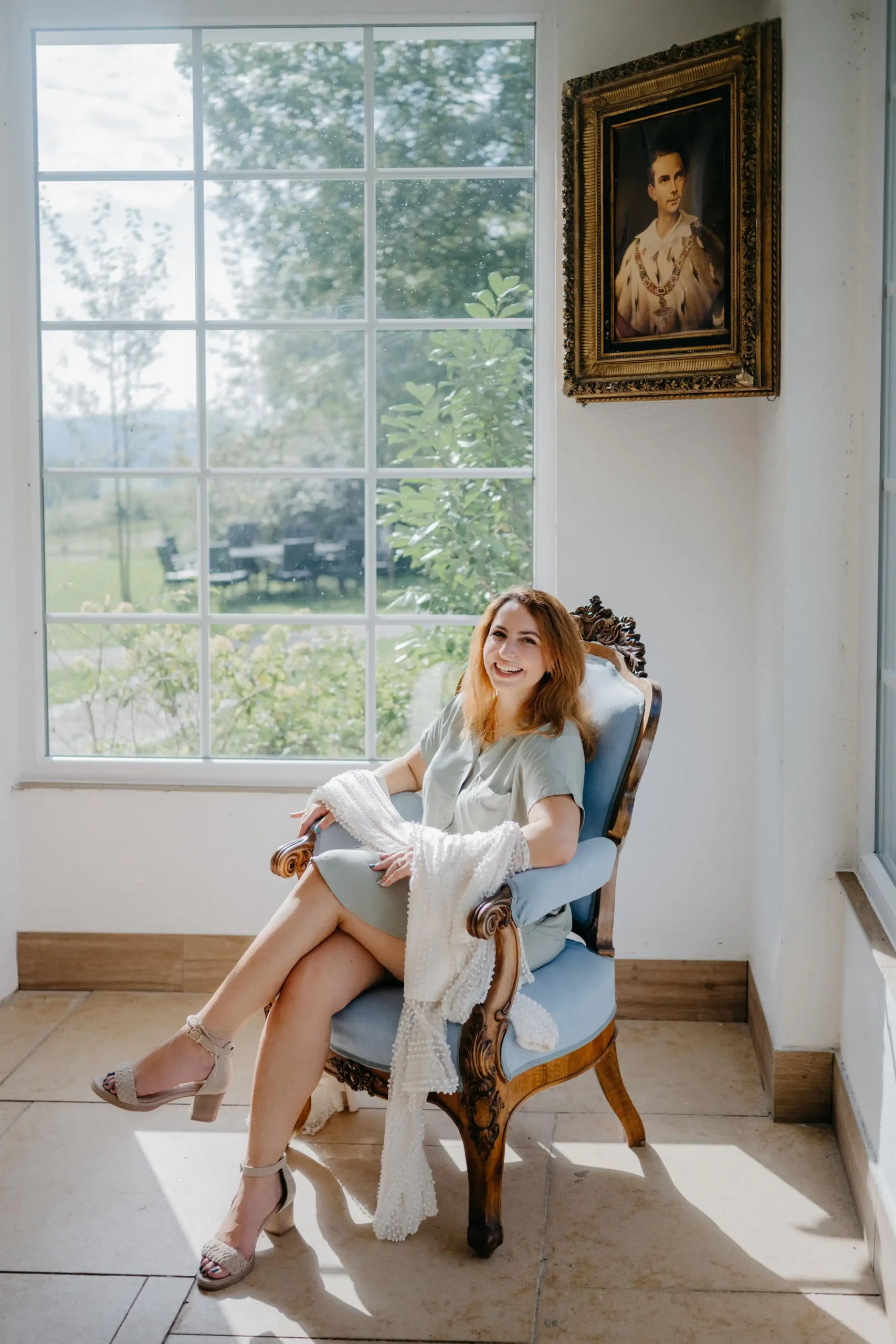 Stefania sitting on a vintage chair by a large window, smiling in natural light.