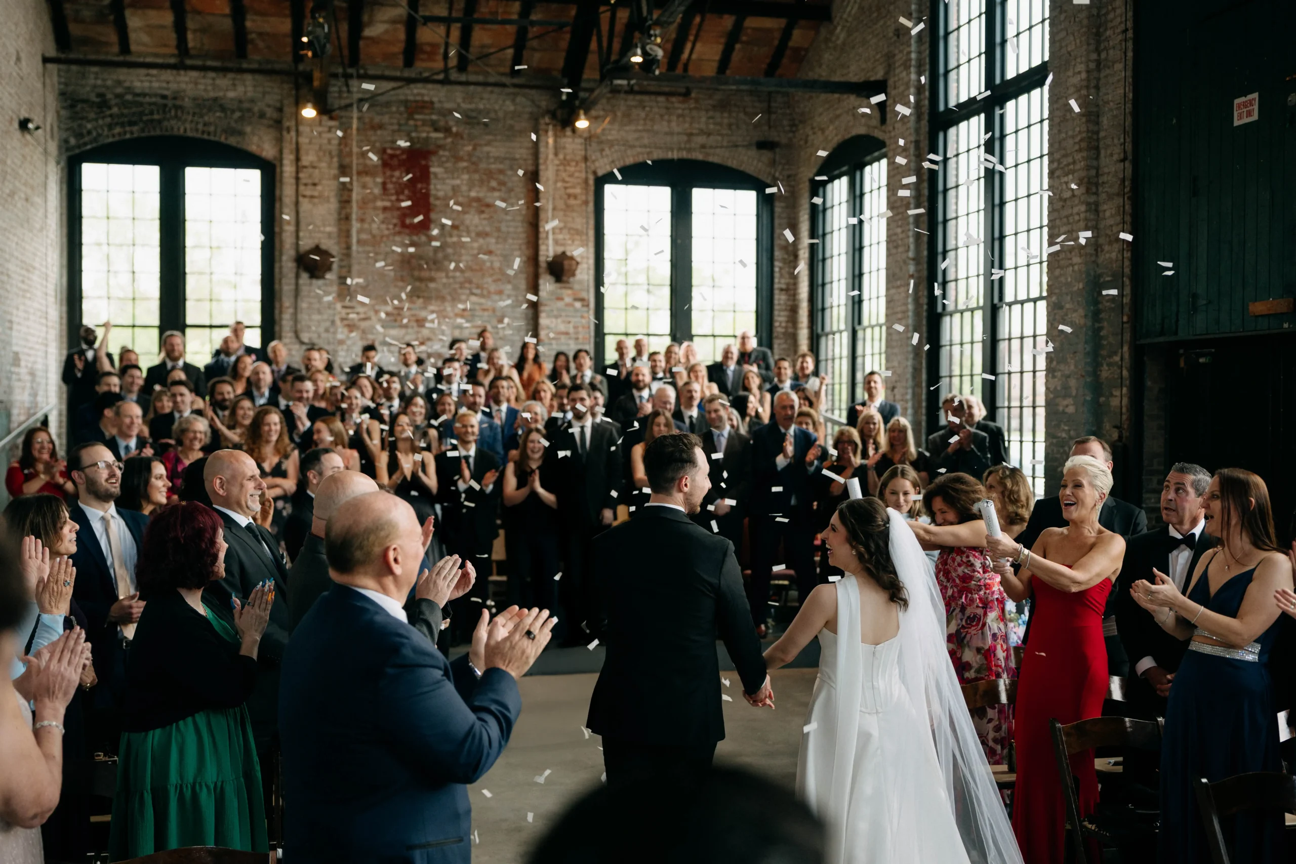 Bride and groom walk down the aisle as guests cheer and throw confetti.