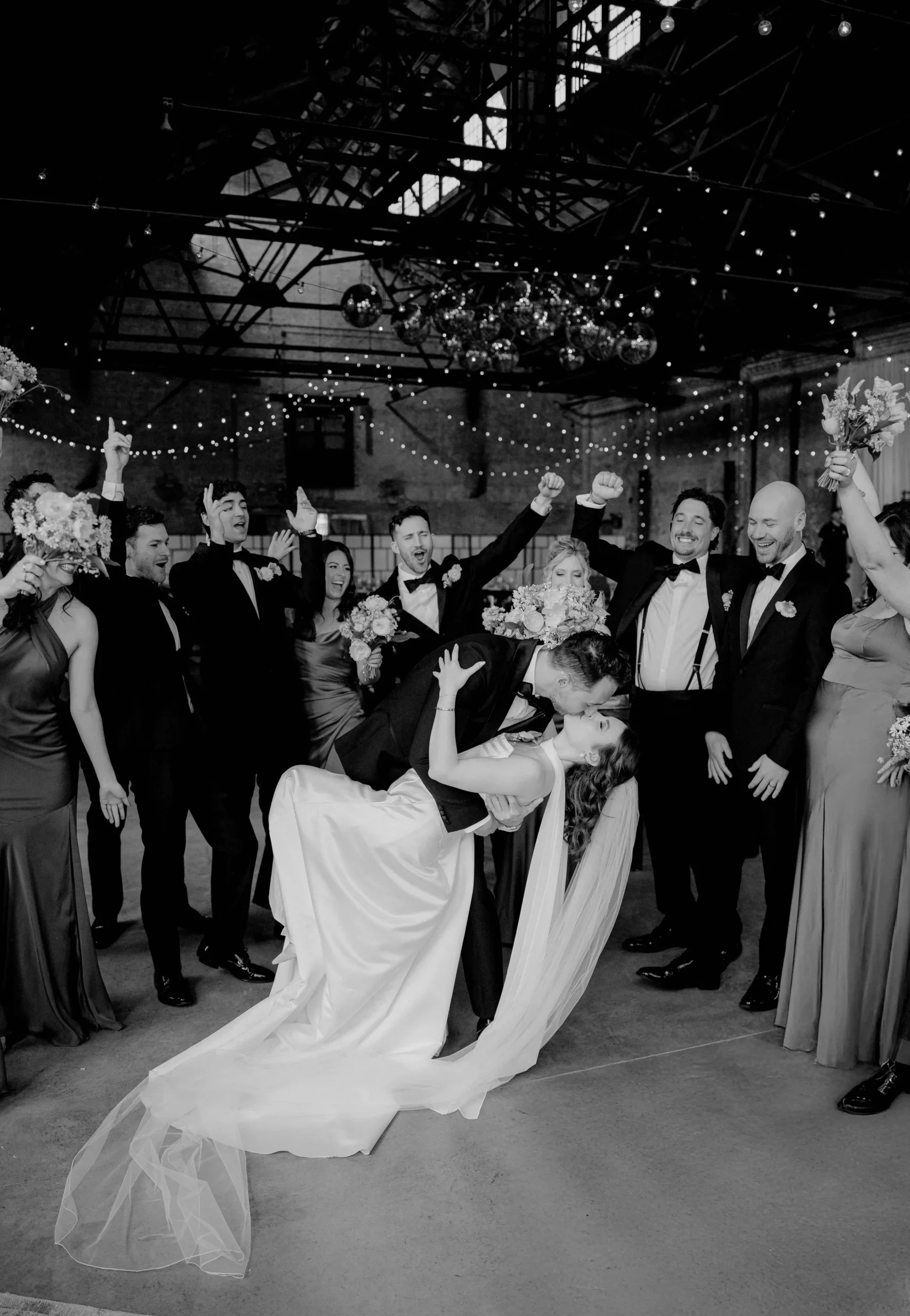 Bride and groom kiss surrounded by cheering guests in an industrial venue.