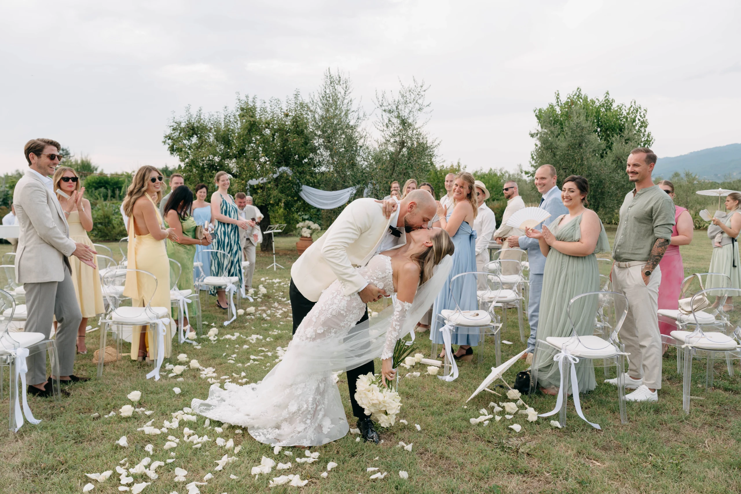 Groom dips the bride in a passionate kiss while guests cheer and applaud during the outdoor ceremony