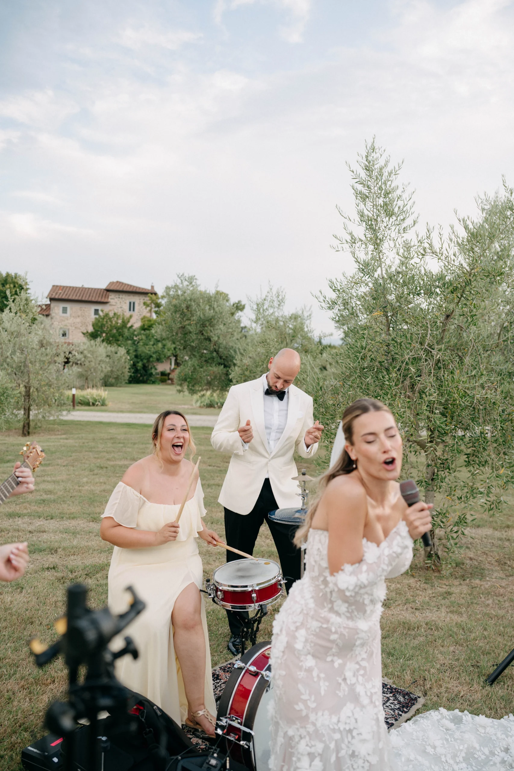Bride sings with mic while groom and friends cheer near a drum set during wedding celebration in an olive grove