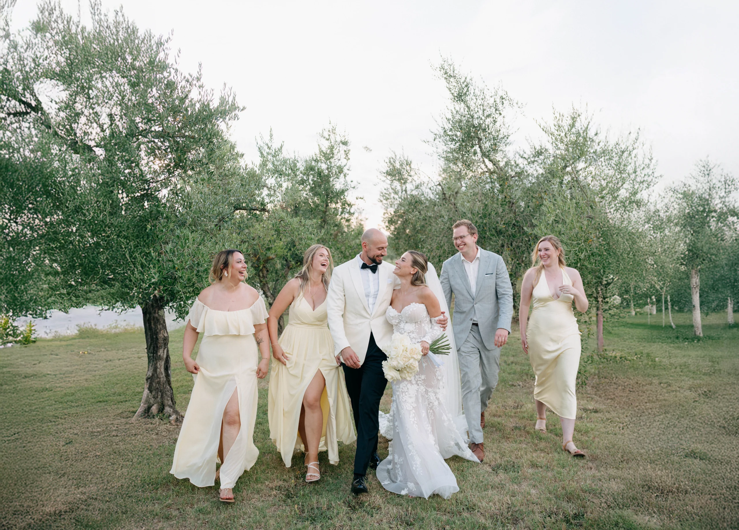 Bride and groom walking happily with bridesmaids and groomsman through olive trees in Tuscany, all smiling