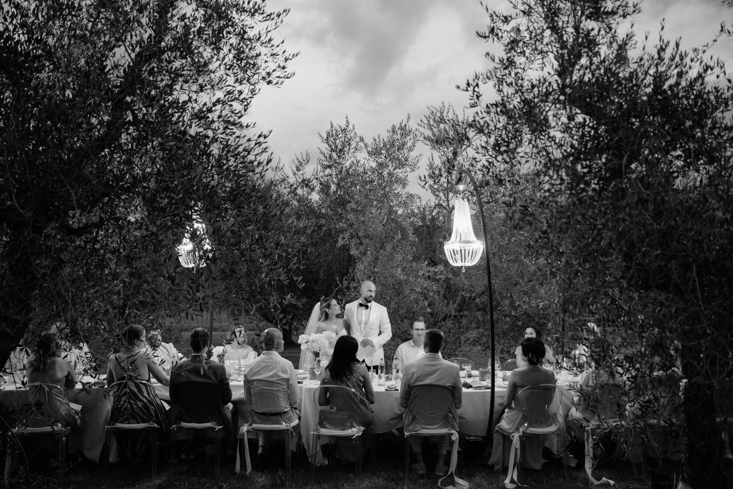 Bride and groom standing at candlelit outdoor dinner table among olive trees during wedding speech in Tuscany