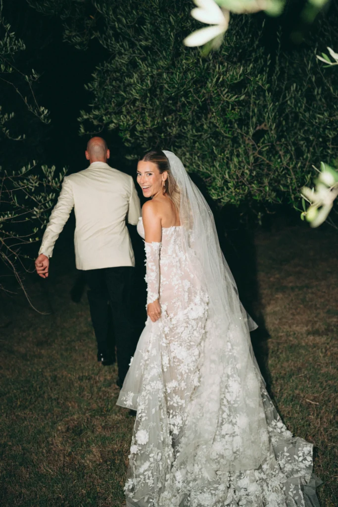 Bride looking back smiling while walking hand in hand with groom at night through olive grove in Tuscany, wearing floral lace wedding dress