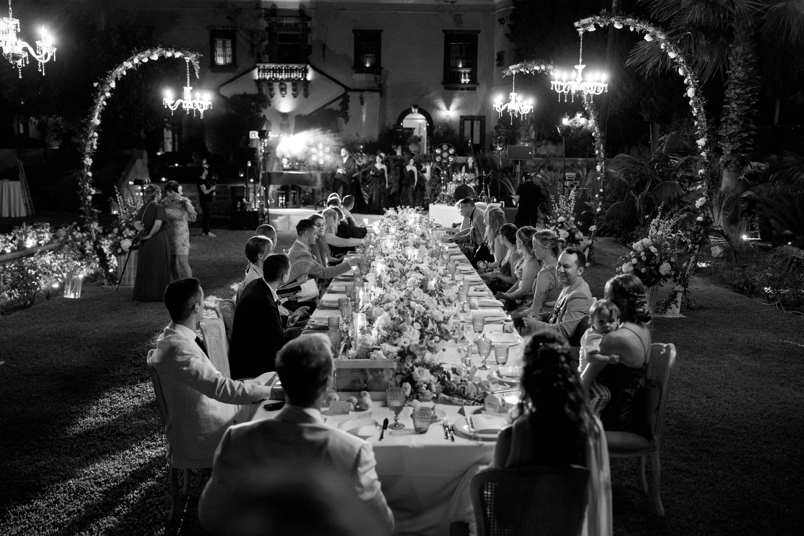 Black and white photo of wedding guests seated at a long dinner table outdoors.