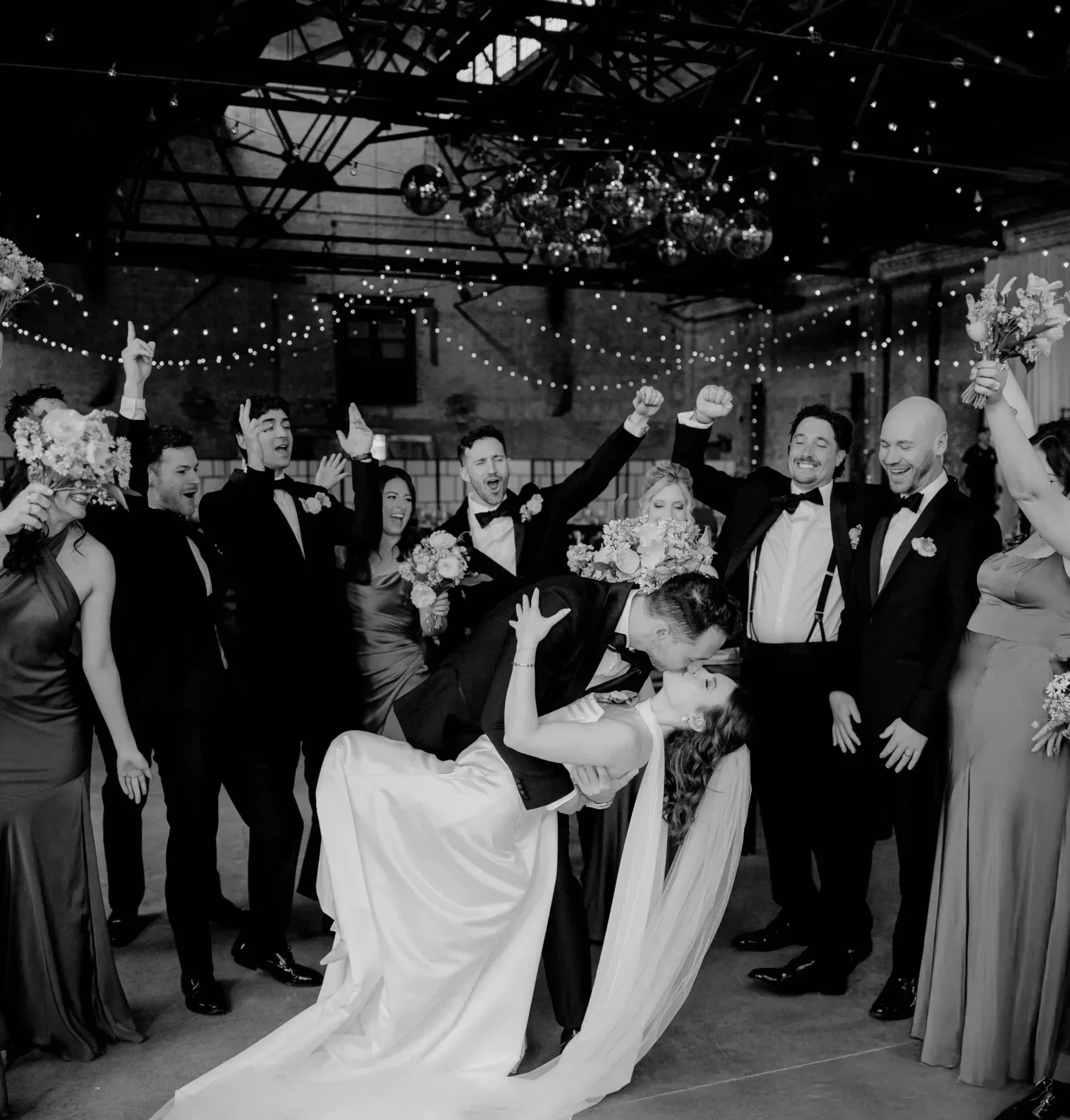 Bride and groom kissing in front of their cheering wedding party at an industrial venue.