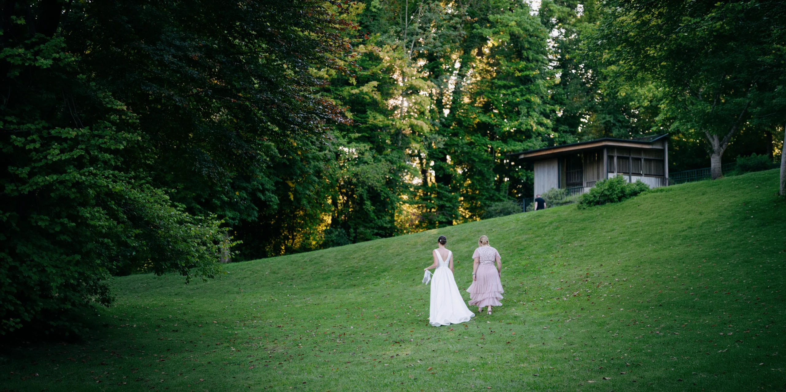 Bride walking with her companion across a wide lawn toward the trees, Munich wedding at La Villa