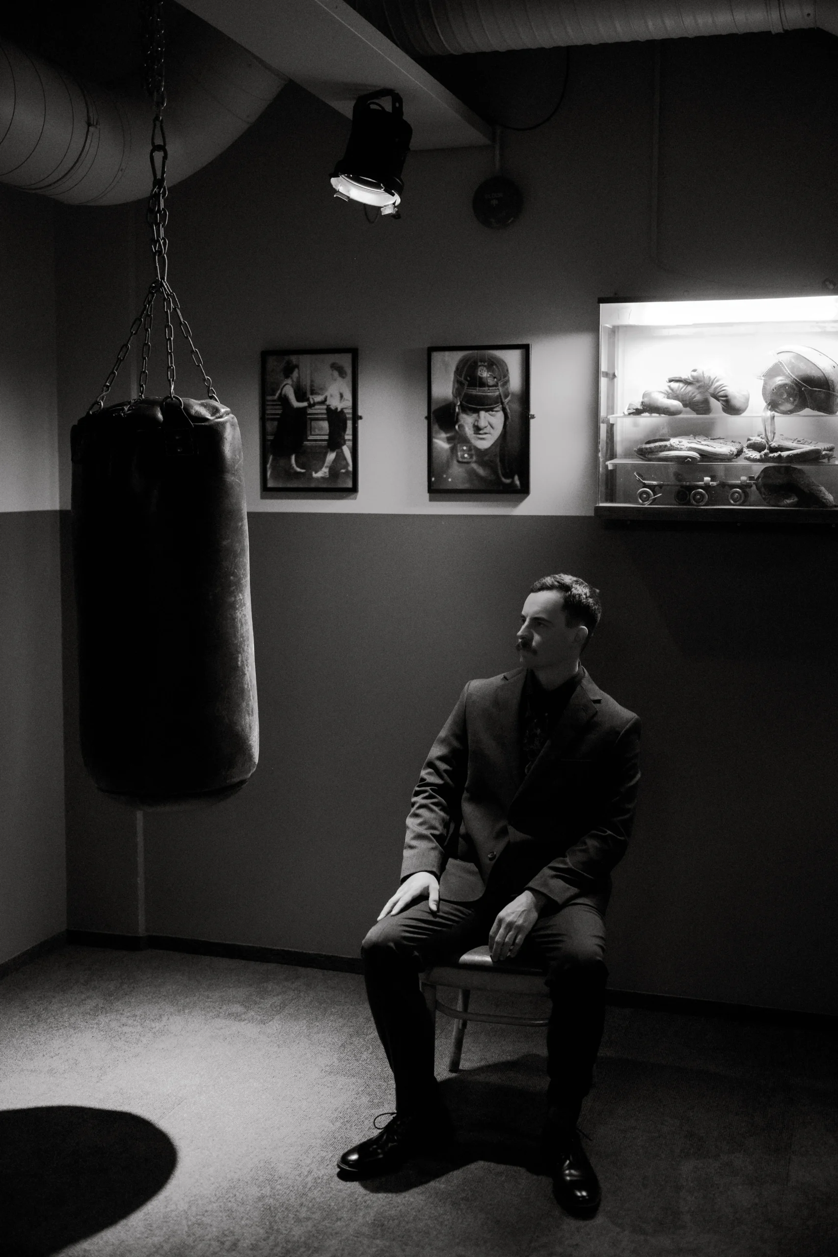 Groom in dark suit sits in moody boxing studio, black and white portrait