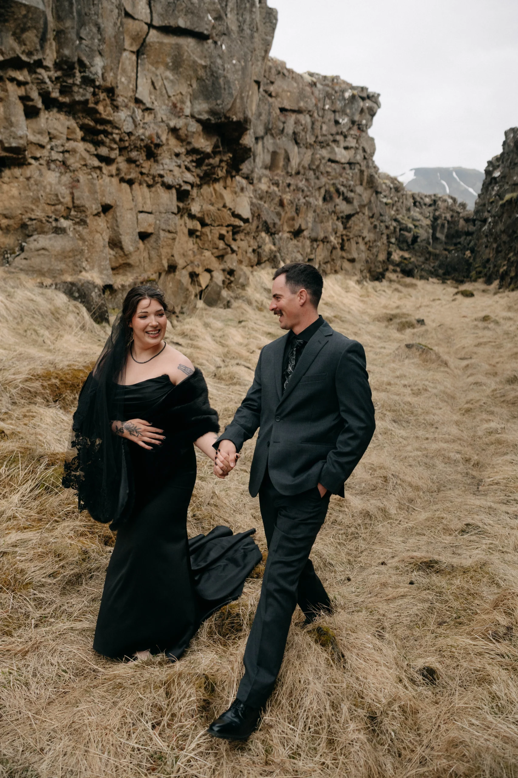 Laughing couple walking through golden grass, bride in black dress