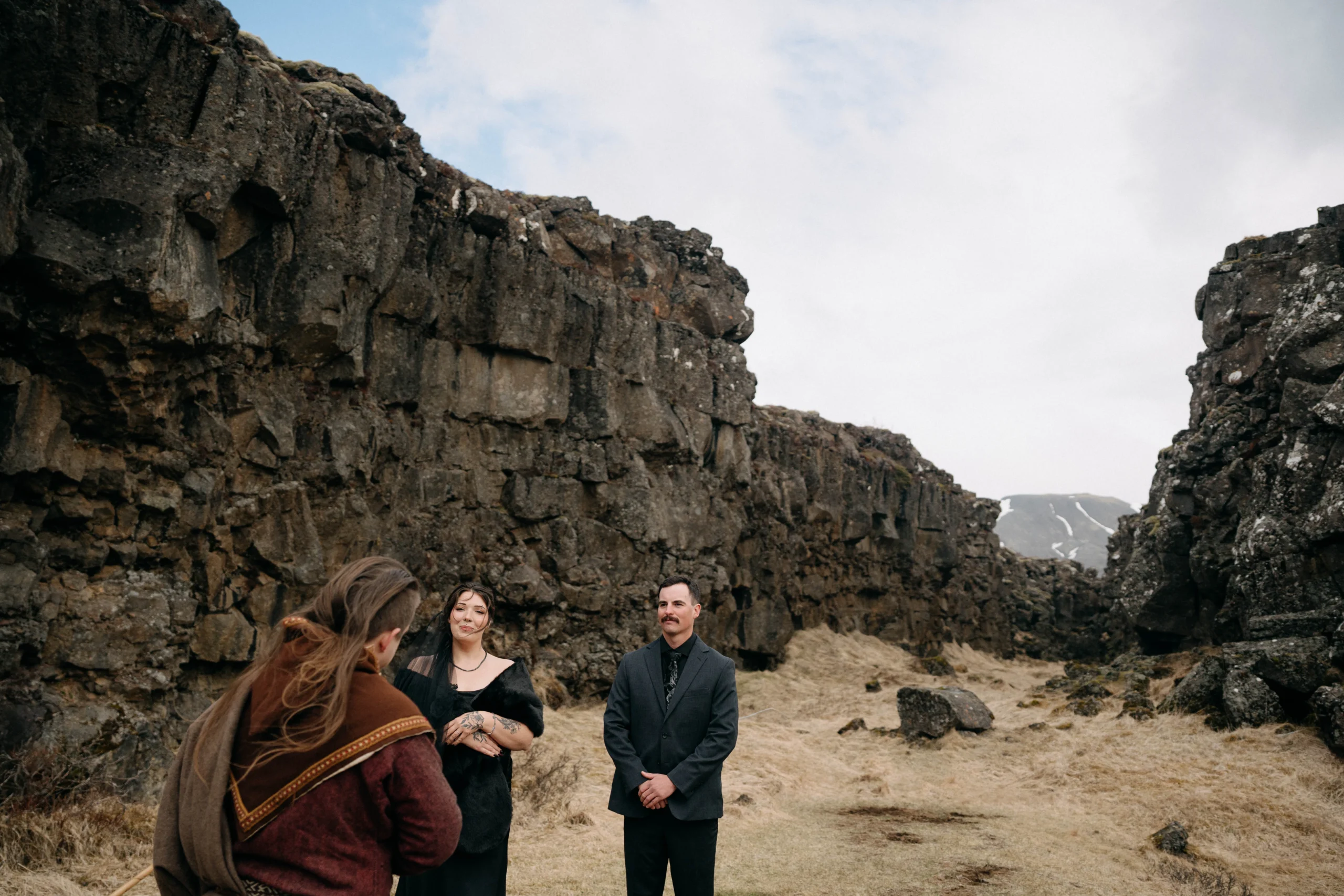 Officiant in historic robes speaking with couple in front of rock wall
