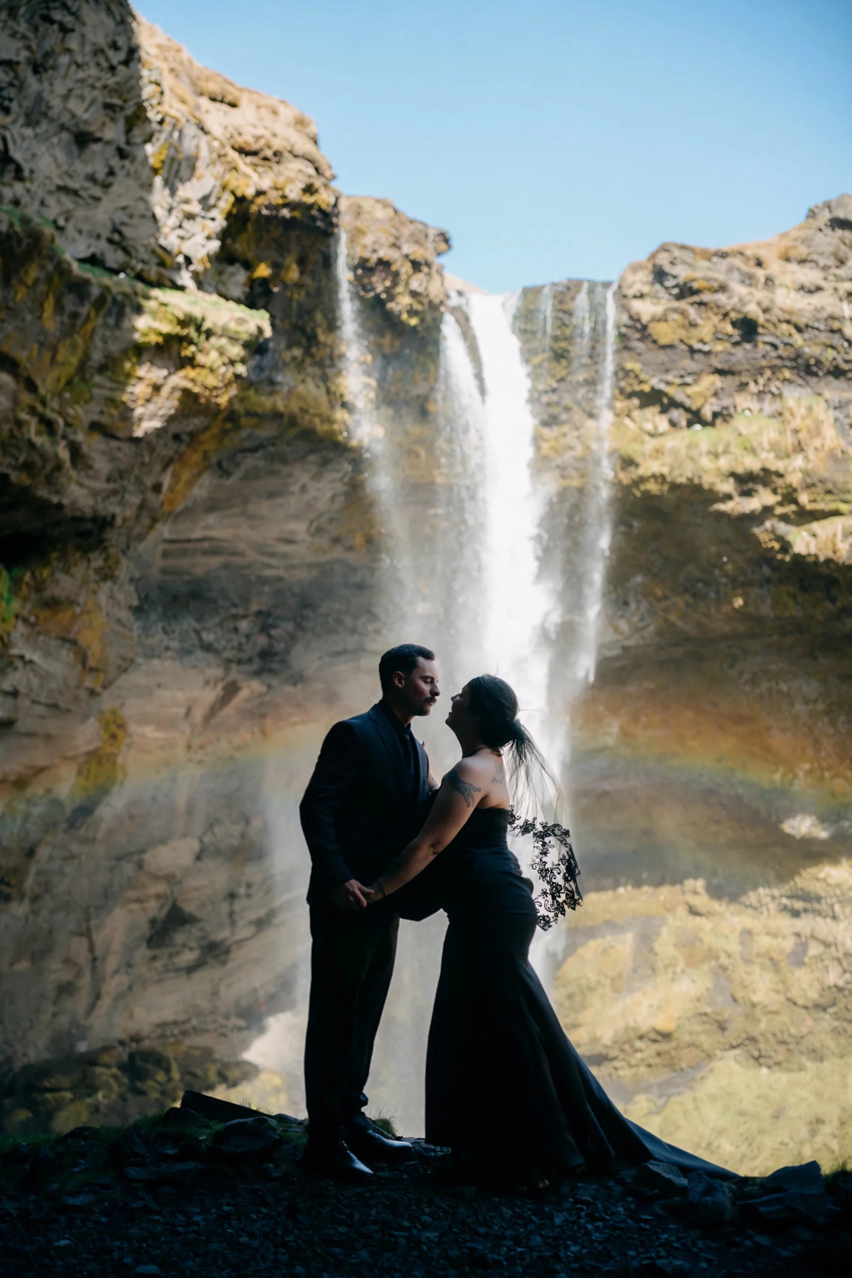Couple in black embrace in front of Icelandic waterfall with rainbow