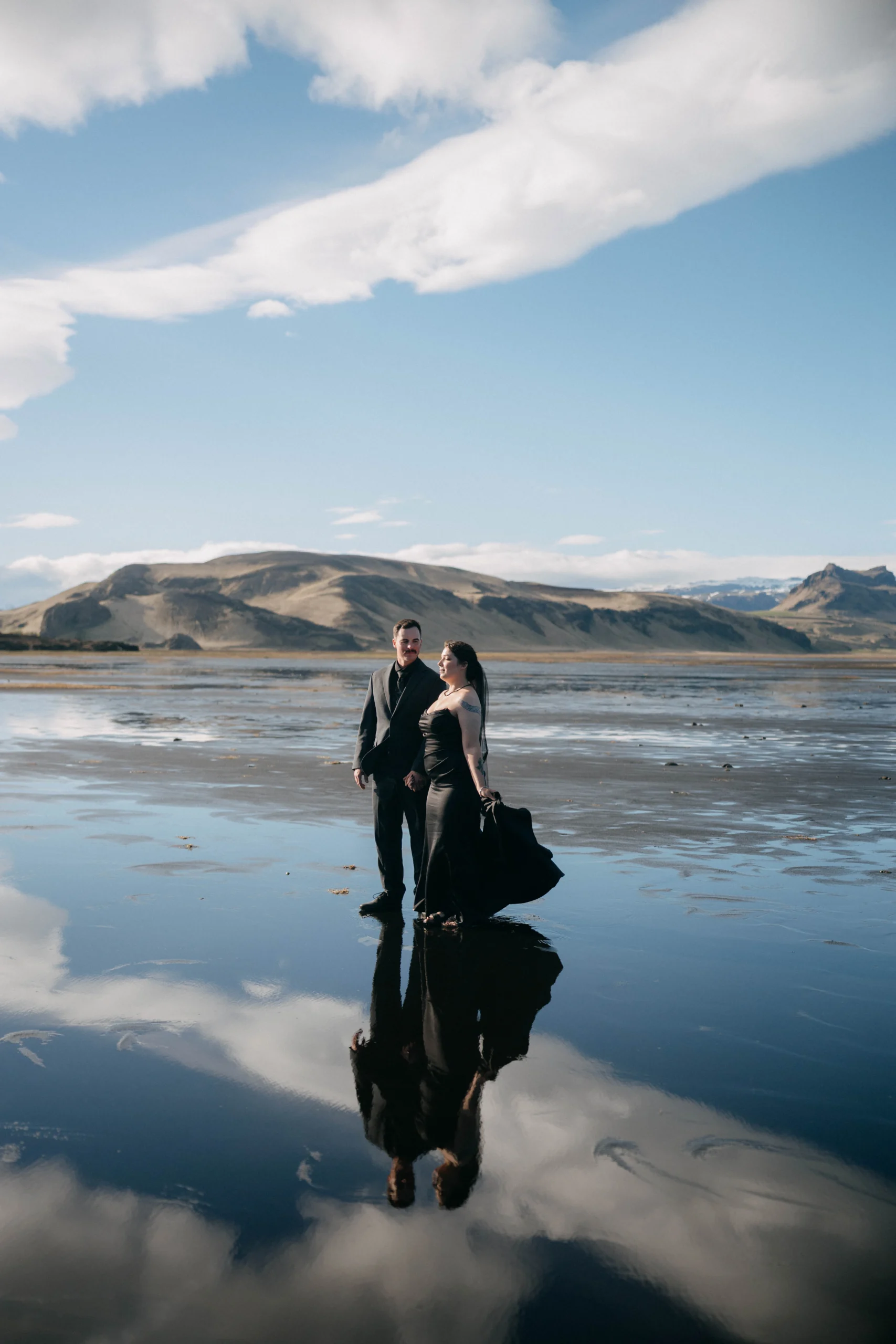 Couple in black at black sand beach in Iceland, mountains reflected in the wet sand at low tide.