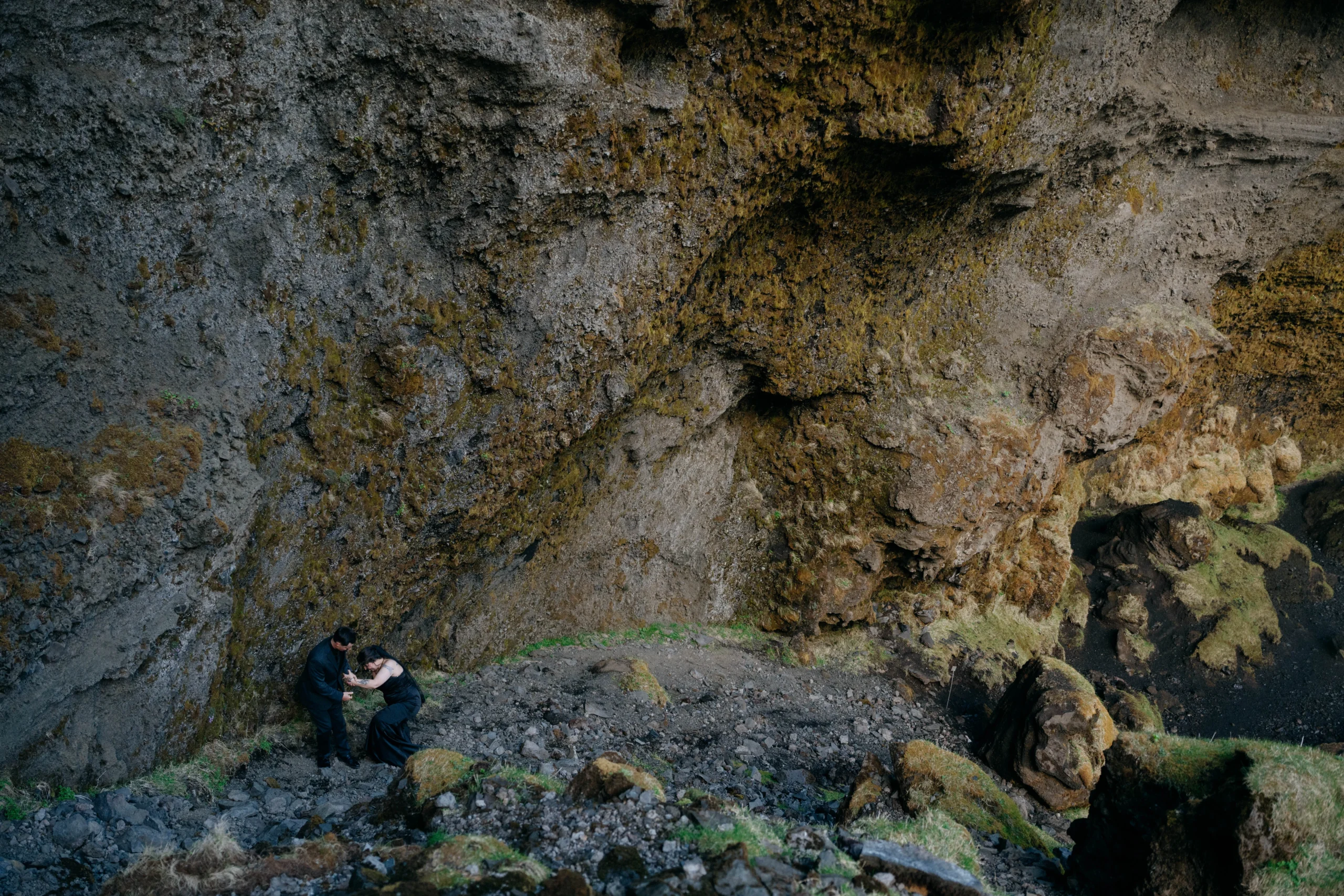 Couple in black attire embracing at the base of a mossy volcanic gorge in Iceland.