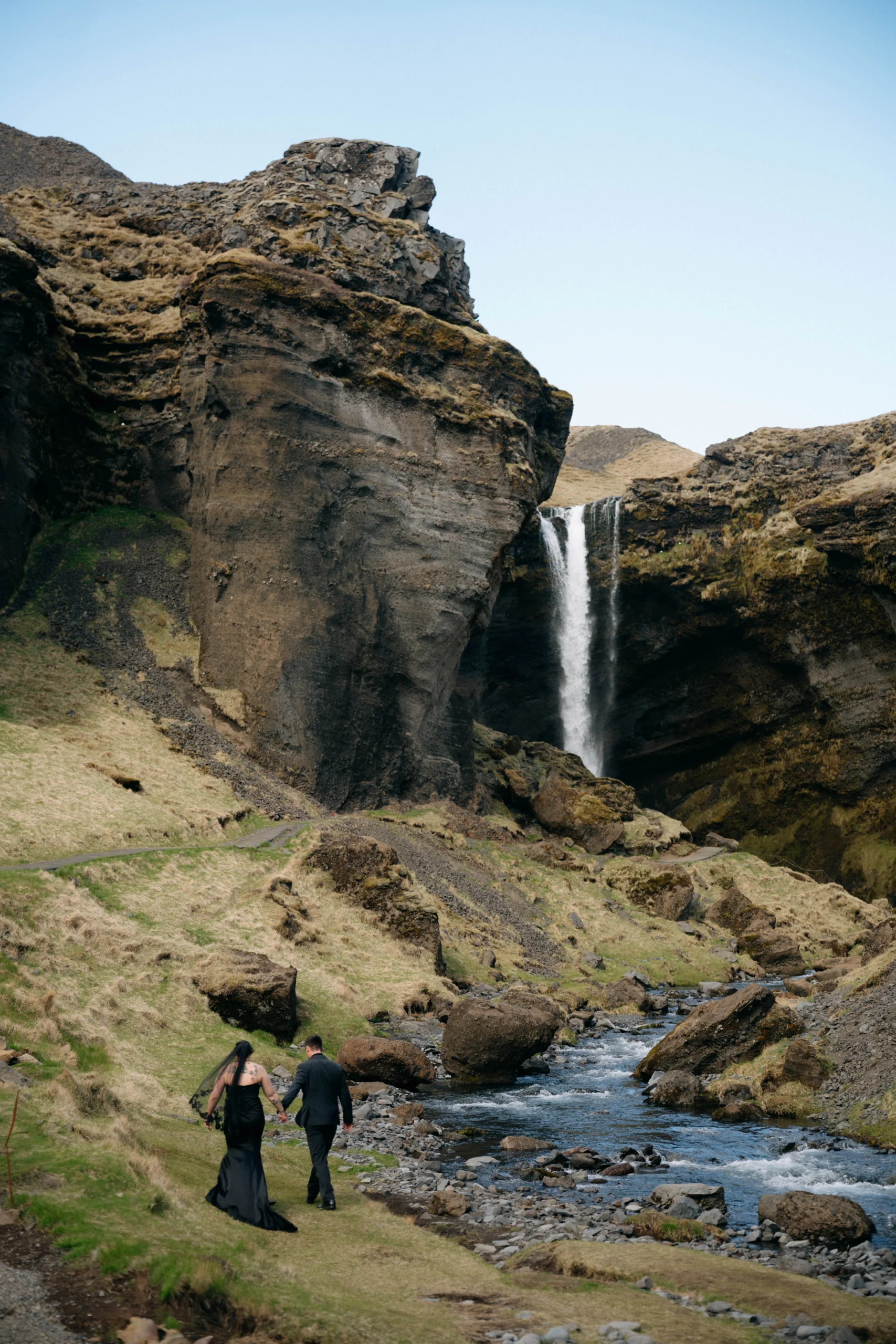 Couple in black wedding attire walking hand in hand along a rocky river toward a distant waterfall in Iceland.