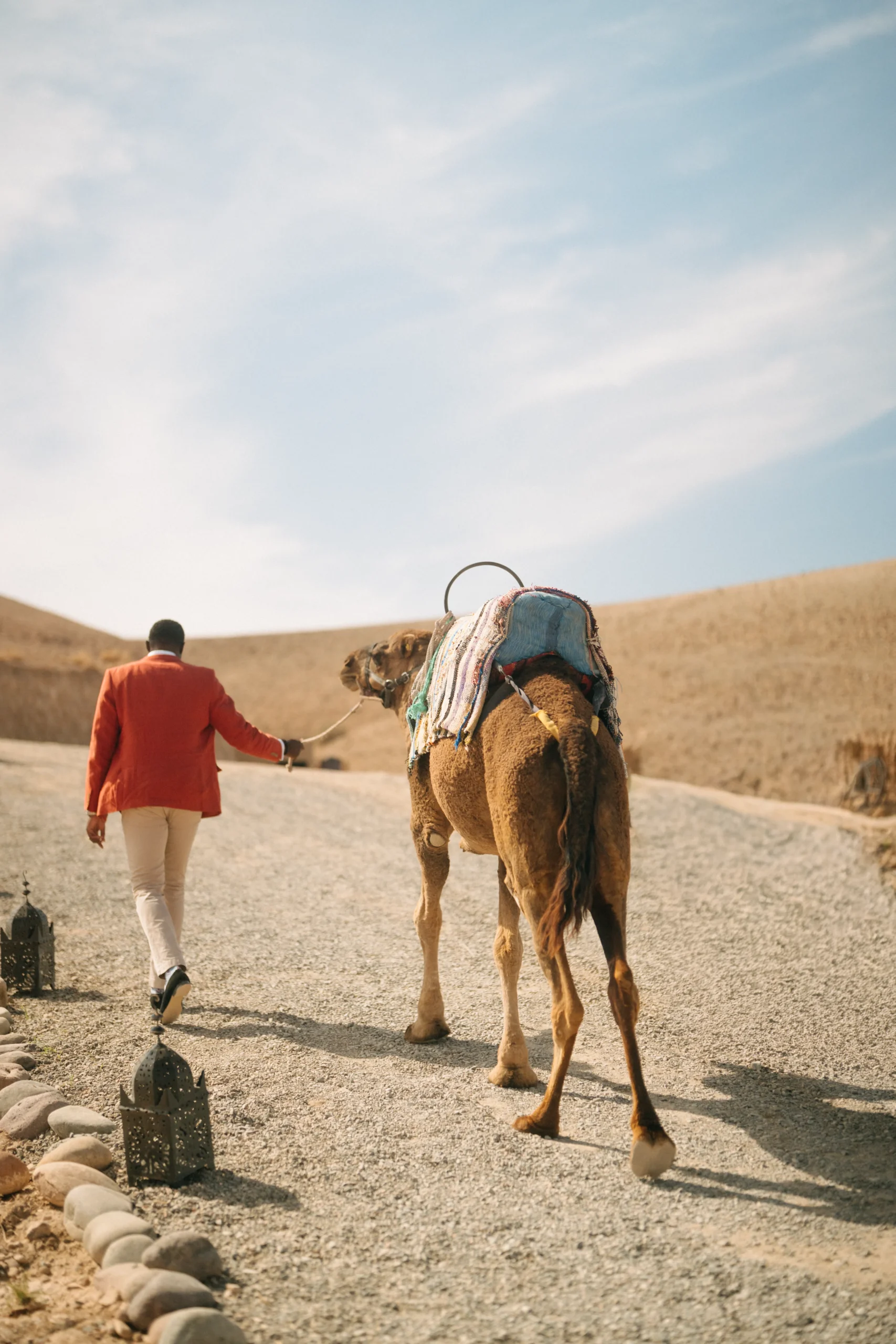 Groom in a red jacket leading a camel along a lantern-lined path at Be Agafay desert camp