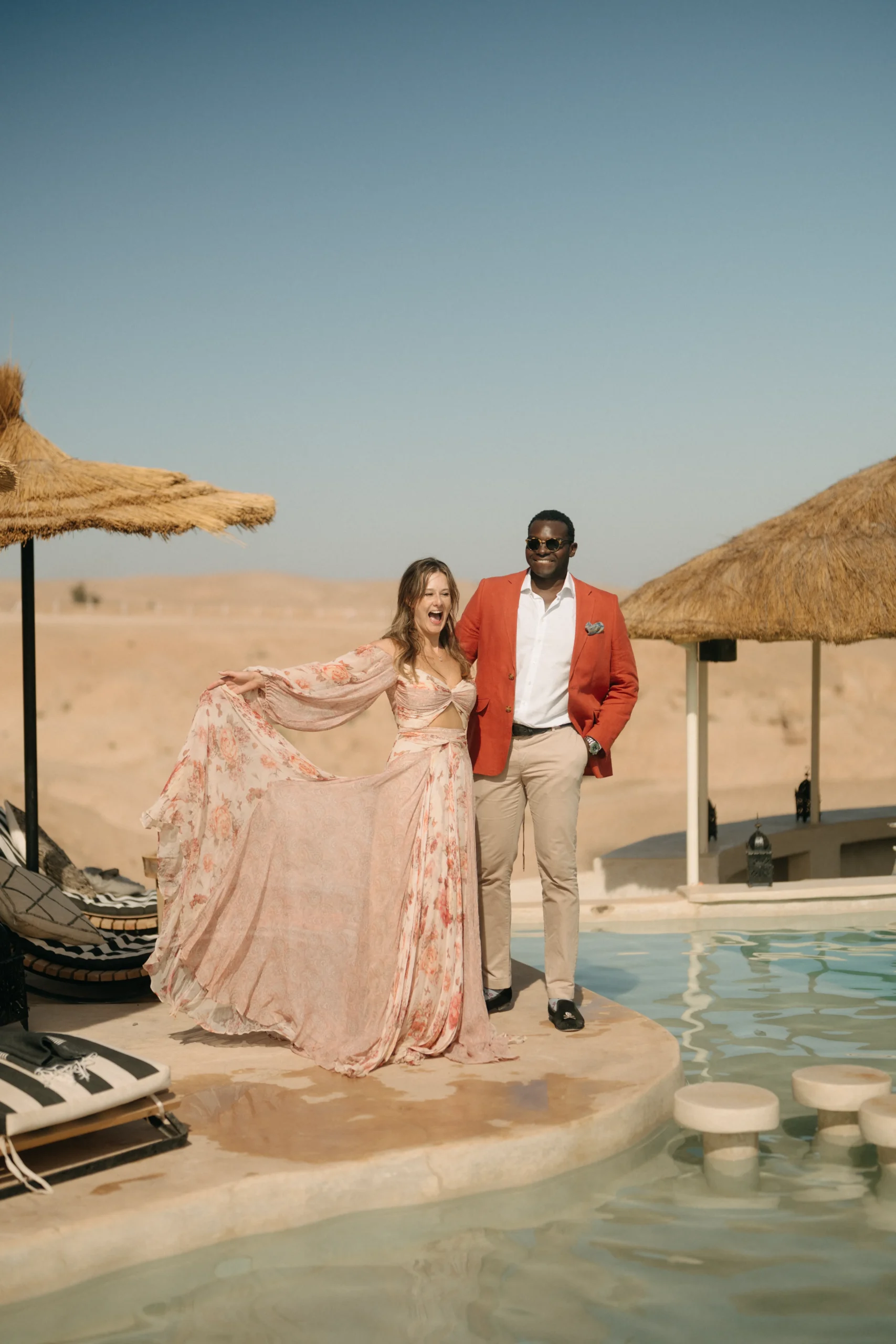 Laughing couple by the pool at Be Agafay desert camp, bride swirling her floral dress